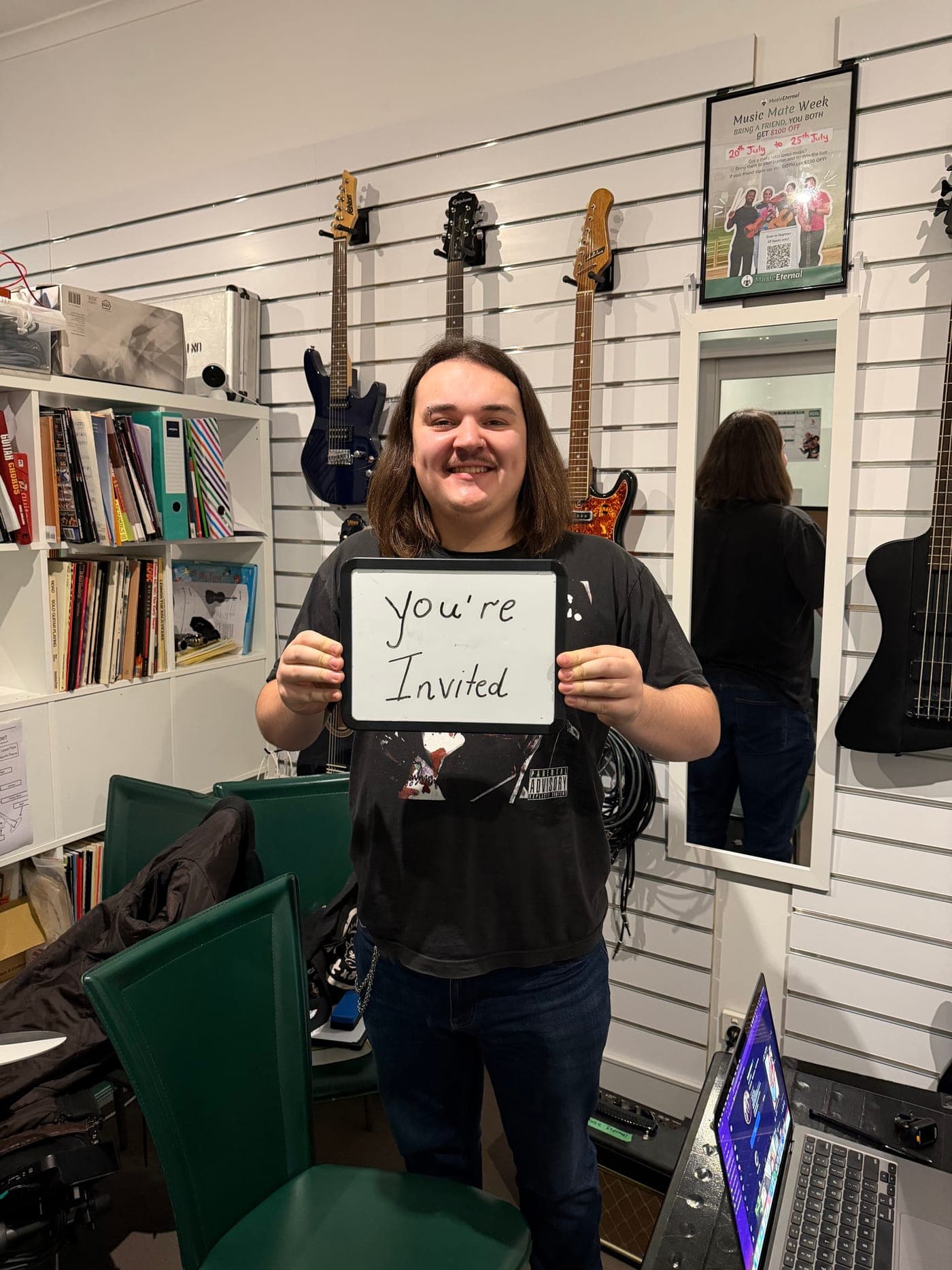 A Music Eternal teacher in front of the guitar wall holding a whiteboard reading: You're invited